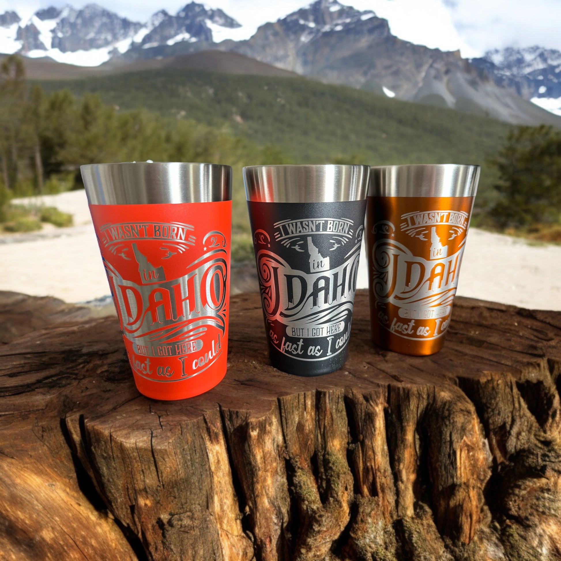 Red, black, and copper engraved Idaho tumblers on a tree stump with Sawtooth Mountains in the background