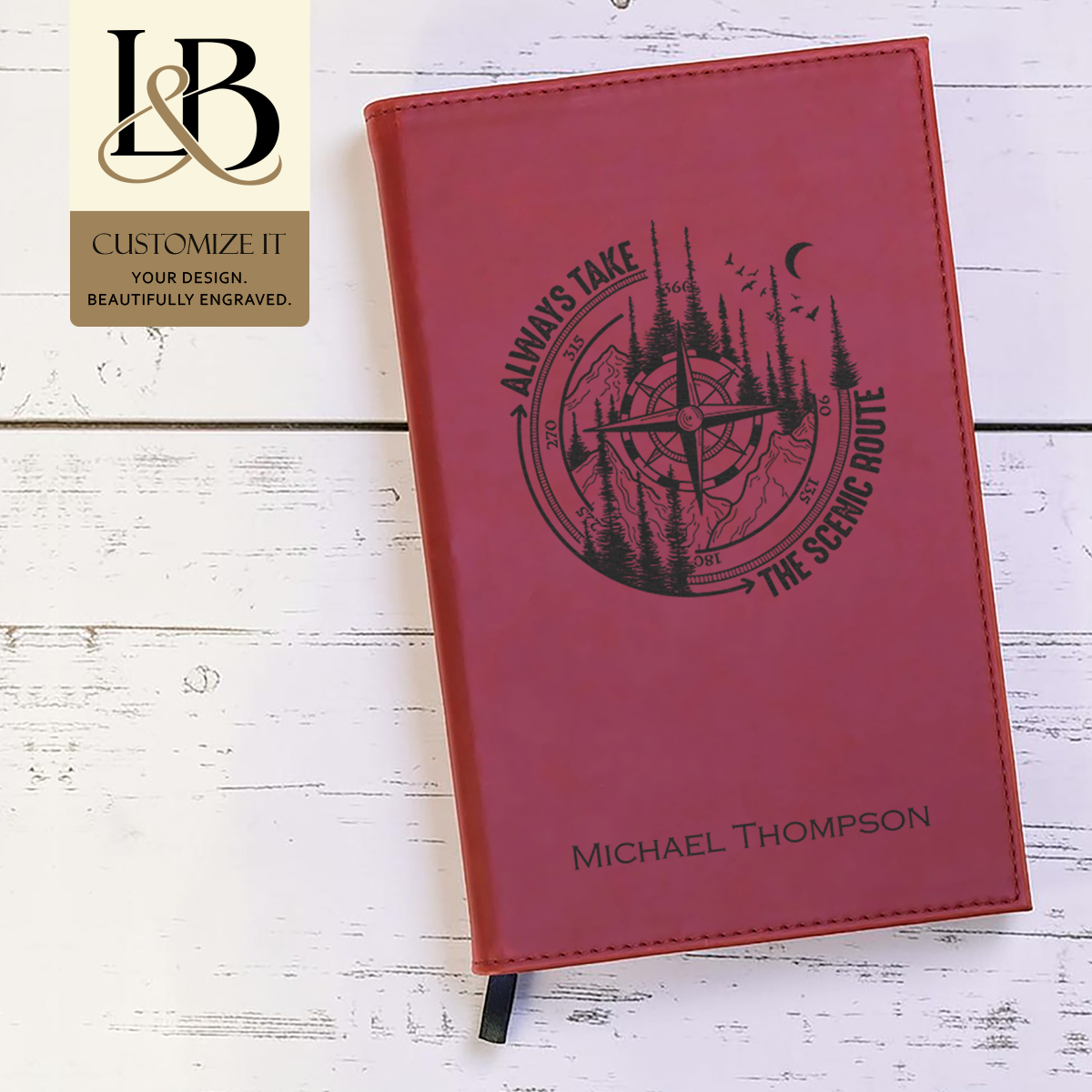 Red personalized notebook with a circular engraved outdoor scene and gold brand badge on pale wood.
