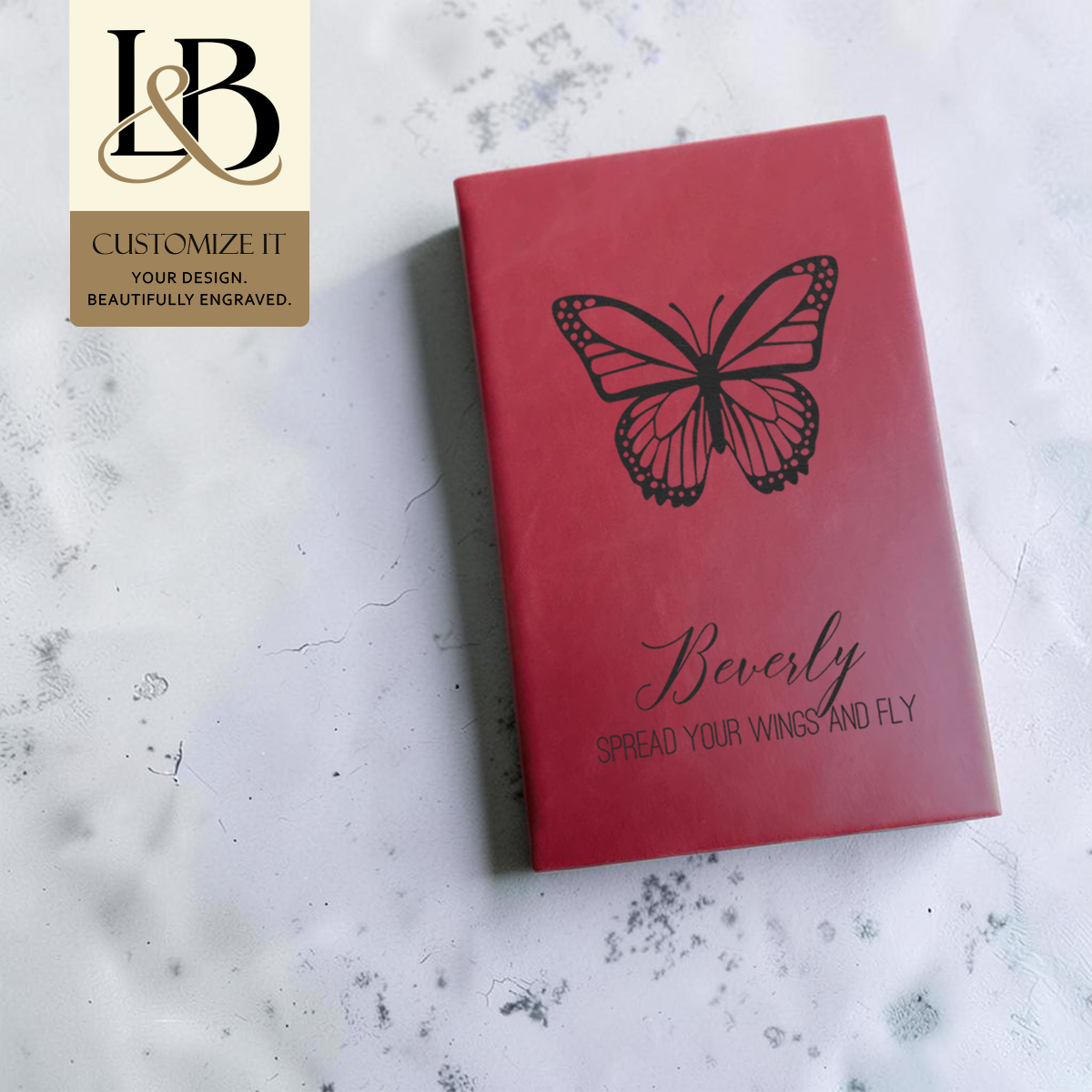 Red personalized notebook with an engraved butterfly and gold brand badge against pale wood.